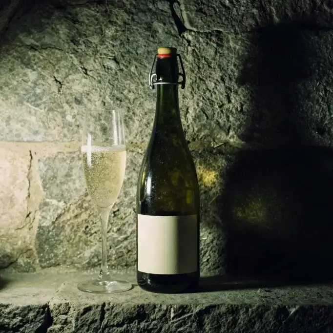 Bouteille de cidre mousseux avec muselet doré dans une cave de vieillissement en pierre, bouteilles couchées sur lattes de bois à l'arrière-plan et flûte remplie de bulles fines.